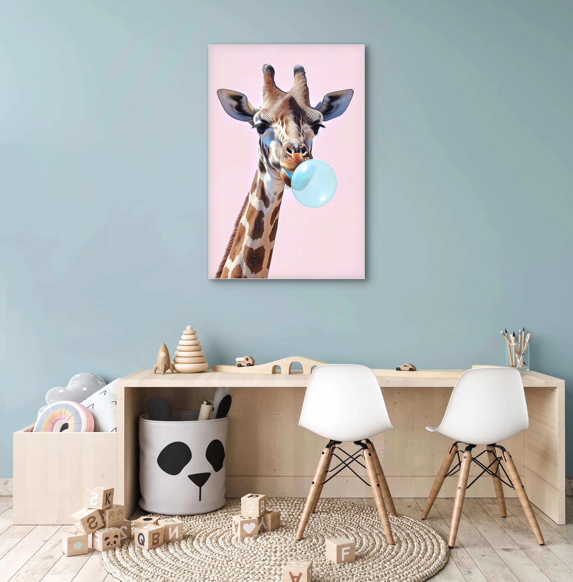 Kunstwerk van een giraffe met kauwgombel aan de muur boven een houten kindertafel met witte stoelen en speelgoed
