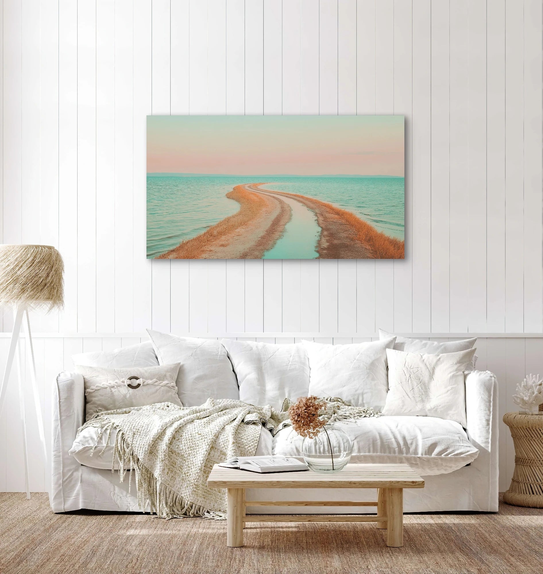 Textielkunstwerk met roze lucht, turquoise zee en zandpad, hangend boven witte bank in een licht interieur
