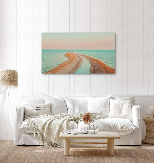 Textielkunstwerk met roze lucht, turquoise zee en zandpad, hangend boven witte bank in een licht interieur
