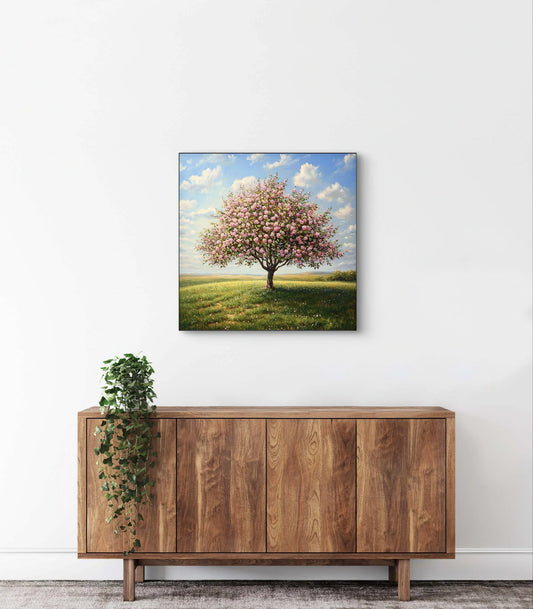 Textielkunstwerk met bloeiende boom in roze tinten onder blauwe lucht, hangend boven een houten kast in een licht interieur
