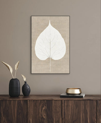 Kunstwerk van een wit blad op beige linnen achtergrond aan de muur boven een donker houten dressoir met decoratie in neutrale tinten
