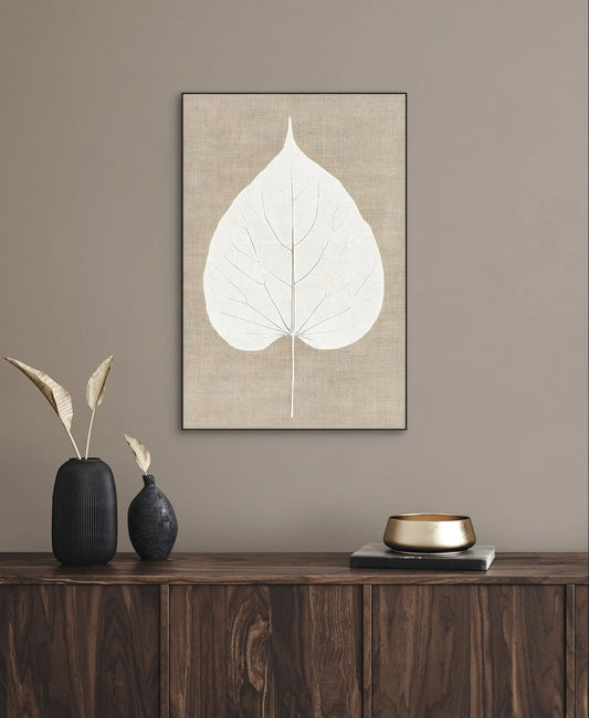 Kunstwerk van een wit blad op beige linnen achtergrond aan de muur boven een donker houten dressoir met decoratie in neutrale tinten
