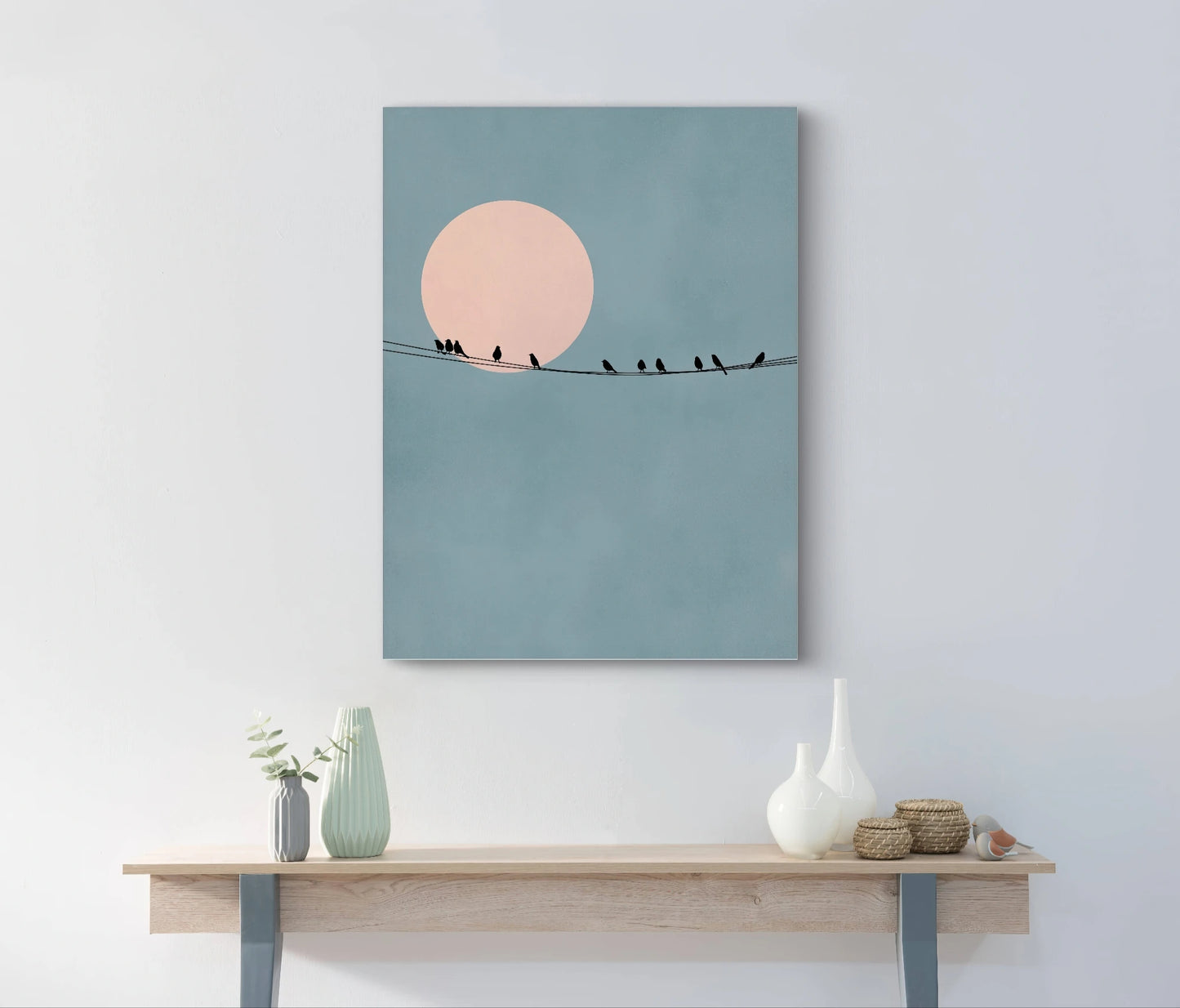 Kunstwerk van silhouetten van vogels op een draad voor een roze zon, aan een muur boven een houten tafel met keramiek in een licht interieur
