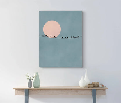 Kunstwerk van silhouetten van vogels op een draad voor een roze zon, aan een muur boven een houten tafel met keramiek in een licht interieur
